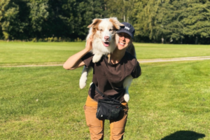 Kvinna står på gräsmatta och har en ljus hund med lång päls på sin rygg. Båda ser glada ut.