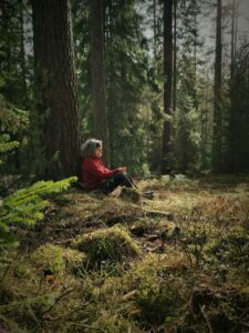 En kvinna sitter i skogen på marken och lutar ryggen mot en tall.