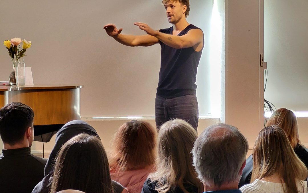 En man med svart linne och grå byxor med kort blont rufsigt hår står på en scen framför en publik. Han håller ut händerna som att han lägger armarna på en osynlig persons axlar.