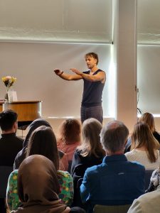En man med svart linne och grå byxor med kort blont rufsigt hår står på en scen framför en publik. Han håller ut händerna som att han lägger armarna på en osynlig persons axlar.