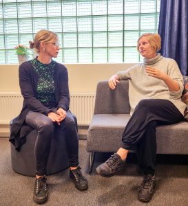 Två kvinnor sitter brevid varandra på en soffa. Den vänstra kvinnan har blont uppsatt hår och glasögon. Klädd i blå kofta grön blommig tröjja och mörka jeans. Den andra kvinnan har ljust hår klippt i page i grå stickad tröja och svarta byxor. De sitte ri samtal med varandra.