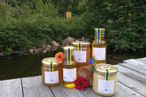 Honungsburkar och flaskor som står i en grupp på ett trädäck, i bakgrunden buskar och vatten.