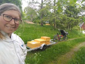 Kvinna med ljus hy och ljust hår med glasögon står i förgrunden med en vit biodlingsrock. I bakgrunden står en cykel med en kärra bakom med ramar på från bikupor.