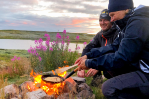 Två personer lagar mat över öppen eld med en sjö och kullar som bakgrund.