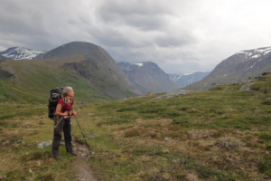 Kvinna med ryggsäck och gångstavar. Står på en stig i grönt landskap och blickar mot en kulle.