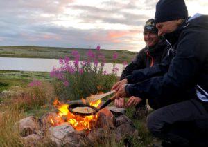 Två personer lagar mat över öppen eld med en sjö och kullar som bakgrund.
