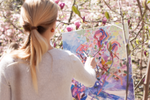 Kvinna står med ansiktet vänt mot en målarduk. Hon målar oljefärg med en pensel, i lila, rosa blå och gult. I bakgrunden ser vi blommande magnoliaträd.