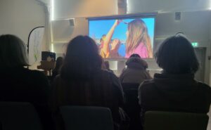 En mörk sal med en bildskärm framför finns siluetter av en publik. På skärmen ser vi en stillbild från en av dansfilmerna. På stillbilden syns det två dansare.