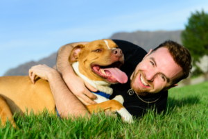 En vit man i medelåldern ligger på gräset och kramar om en ljusbrun hund av rasen staffordshire terrier.